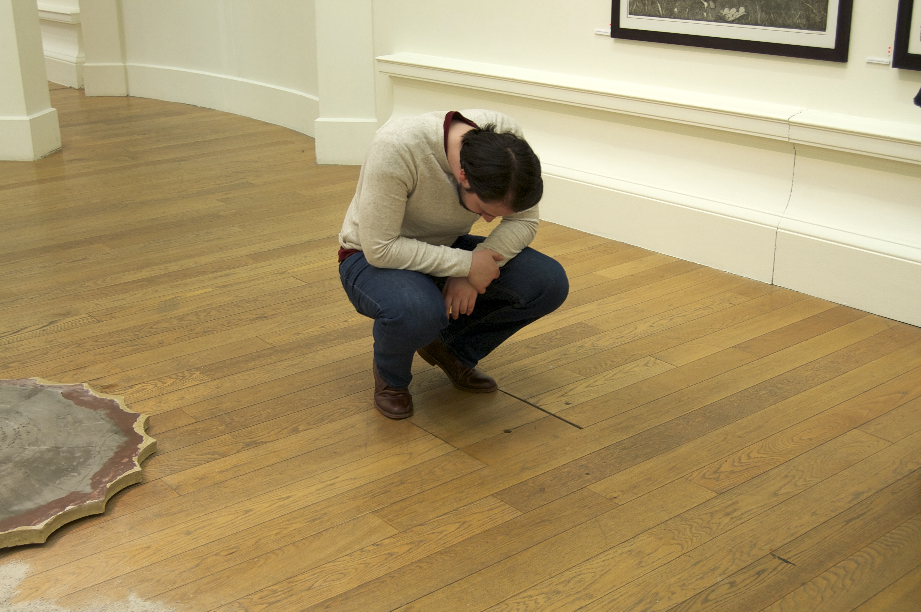 A gallery space with wooden floors. A man crouches down to look into a small hole in the floor.