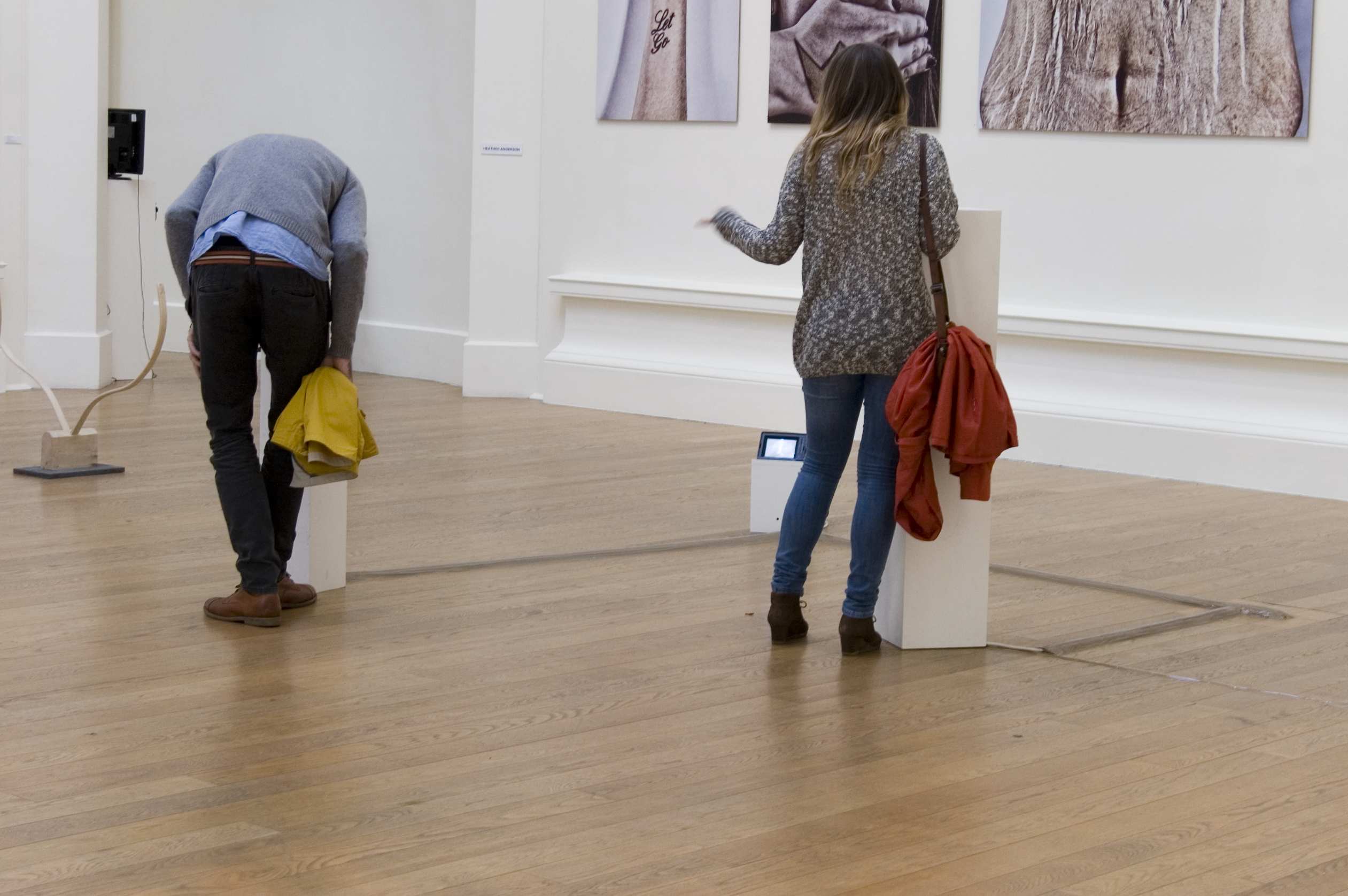 A gallery space. Two white plinths are on the floor, a couple of metres apart. A man is looking into one of them, while a woman stands beside the second, she is pointing at the man.